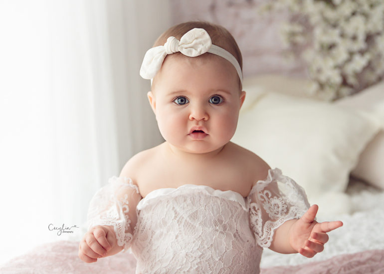 a baby girl in white dress sitting on the bed with smiling to the camera